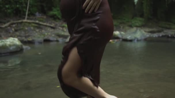 Krásná těhotná žena v tmavě hnědých šatech se na vodě v tropické džungli nachází bosý. Zpomaleně. — Stock video