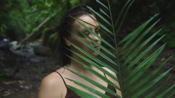 Retrato de una hermosa mujer embarazada con una enorme hoja de palma en sus manos junto a un río tropical en la selva. Movimiento lento . — Vídeo de stock