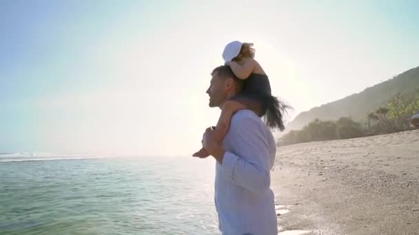 어깨에 앉아 있는 딸을 가진 아버지가 바다 해변 근처를 걷고 있습니다.. — 비디오