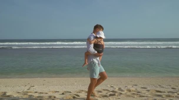 Счастливый отец держит в руках милую дочь и гуляет по пляжу в солнечный день — стоковое видео
