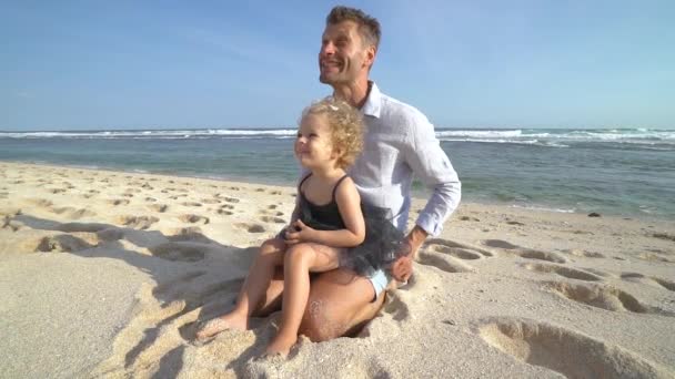 Счастливый отец с дочерью на коленях веселятся на пляже в солнечный день — стоковое видео