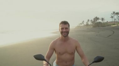 Gün batımında siyah kum sahilde bir motosiklet üzerinde oturan mutlu yakışıklı adam