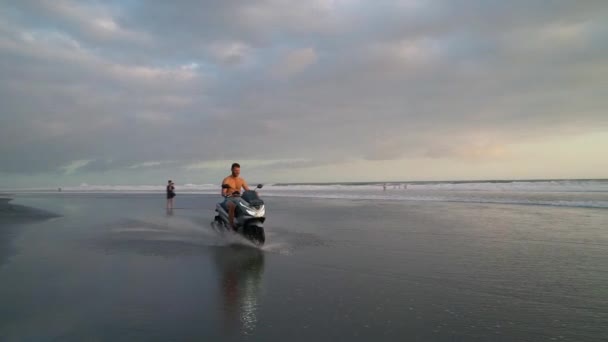 Uomo felice cavalcando in moto sulla spiaggia di sabbia nera e acqua di mare al tramonto — Video Stock