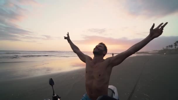 Joyeux bel homme assis sur une moto sur la plage de sable noir au coucher du soleil — Video