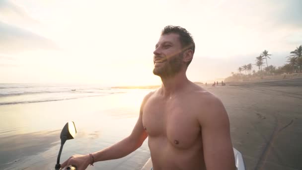 Lycklig stilig man sitter på en motorcykel på den svarta sandstranden vid solnedgången — Stockvideo