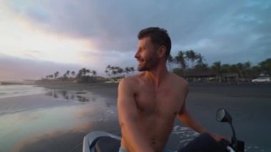 Gün batımında siyah kum sahilde bir motosiklet üzerinde oturan mutlu yakışıklı adam