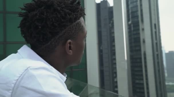 Primer plano del joven africano guapo en camisa blanca mira a la ciudad desde el piso alto de un rascacielos — Vídeos de Stock