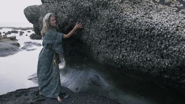 Молода жінка з пасмами натурального сірого волосся на фоні каменю, покритого мушлями біля океану — стокове відео