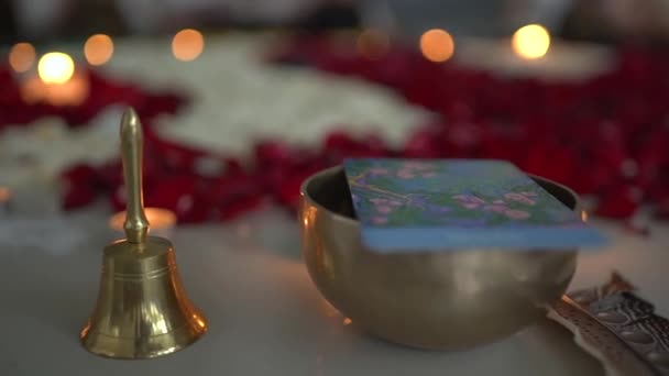 Primer plano de un cuenco de canto tibetano, timbre, cartas del tarot sobre el fondo de pétalos de rosa y velas ardiendo durante una práctica espiritual femenina . — Vídeo de stock