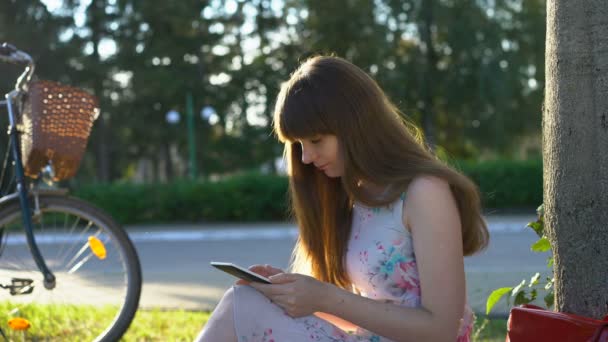 Mujer romántica joven sentada en el parque, leyendo e-book, sonriendo a sí misma. Verano — Vídeo de stock