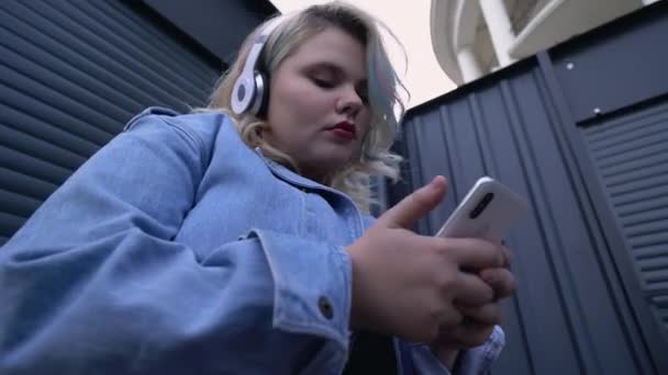 Mujer joven con estilo escuchando música en los auriculares, utilizando la aplicación móvil en el teléfono inteligente — Vídeo de stock