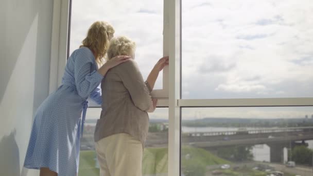 Mujer rubia abrazando a madre, mirando a la ciudad a través de la ventana, juntos — Vídeo de stock