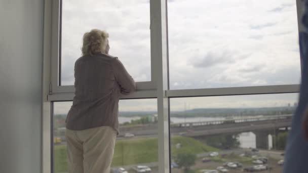 Hija cariñosa abrazando a madre anciana, de pie cerca de la ventana, mirando a la ciudad — Vídeo de stock
