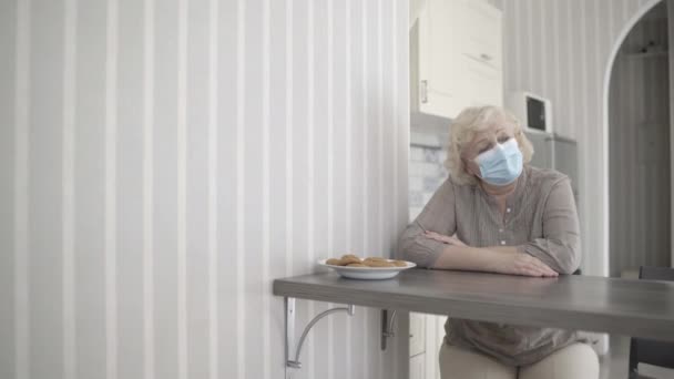 高齢者の女性は、医療用マスクを着て、キッチンテーブルに座って、自己分離 — ストック動画