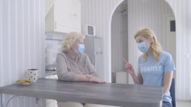 Korunmak için yüz maskesi takan yaşlı bir kadını evinde ziyaret eden gönüllü bir kadın.