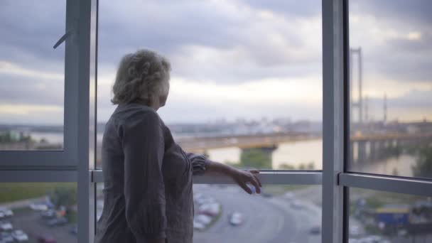 Ältere Frau steht in der Abenddämmerung am Fenster und blickt auf den hektischen Stadtverkehr, einsam — Stockvideo