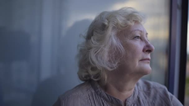 Old wrinkled woman standing by window alone missing family, suffering depression — Stock Video