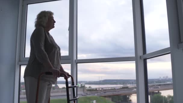 Old woman standing by window with walking frame, rehabilitation after surgery — Stock Video