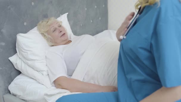 Больная старуха лежит в постели, разговаривает с молодым врачом, медицинские услуги на дому — стоковое видео
