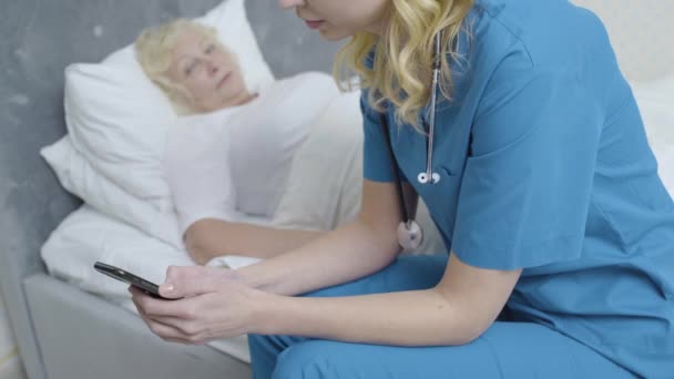 Грубый ухаживающий прокручивает смартфон, игнорирует больного старого пациента, некомпетентность — стоковое видео