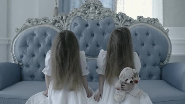Dos niñas misteriosas sentadas en un sofá vintage, sosteniendo un juguete sangriento, horror — Vídeos de Stock