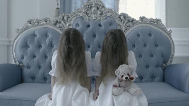 Creepy twins staring at cam sitting on old sofa with bloody toy in hands, zombie — Stock Video