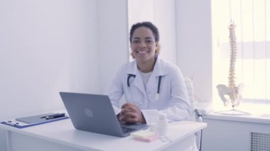 Kamerada gülümseyen kadın doktor, masada oturuyor, tıbbi servis, iş.