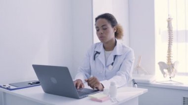 Laptop üzerinde çalışan Afro-Amerikalı kadın doktor test sonuçlarını analiz ediyor.