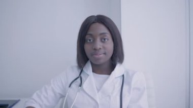 Steteskoplu Afro-Amerikalı doktor kameraya, tıbbi personele, kliniğe bakıyor.