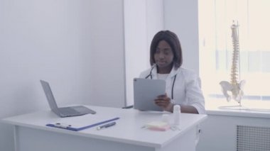 Afro-Amerikalı terapist internetten hastanın analizini kontrol ediyor, iş yerinde tablet kullanıyor.