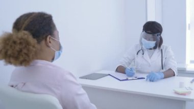 Koruyucu yüz rozeti takan Afro-Amerikalı doktor hastanın belirtilerini yazıyor.