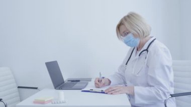 Maskeli pratisyen doktor ofis masasında rapor hazırlıyor.