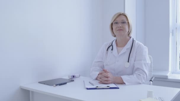 Médico profesional sonriendo en cámara, sentado en la mesa en la clínica, el trabajo — Vídeos de Stock