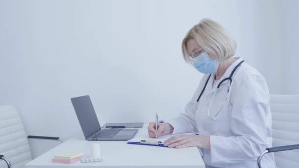 사무실 탁자에서 마스크를 쓰고 있는 의료인이 보고서를 준비하는 모습 — 비디오