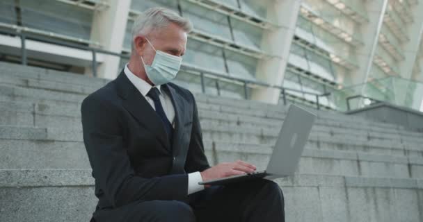 Yüz maskeli bir iş adamı dışarıda dizüstü bilgisayarda çalışıyor, salgın sırasında iş yapıyor. — Stok video