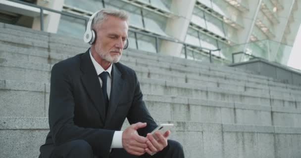 Розслаблений бізнесмен слухає музику в навушниках, використовуючи мобільний додаток на гаджеті — стокове відео