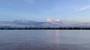 Gün batımında Mekong Nehri 'nin sakin suları pastel gökyüzünü ve manzaralı bir dağ sırası tarafından çerçevelenmiş parlayan bulutları yansıtıyor. Seyahat, doğa ve arka plan kullanımı için ideal sakin bir manzara.