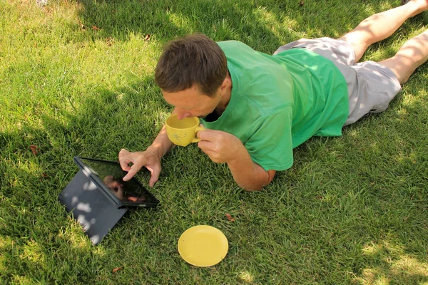 Orta yaşlı bir adam çimenlerin üzerinde yatıyor, çay içiyor ve tablet bilgisayara bakıyor..
