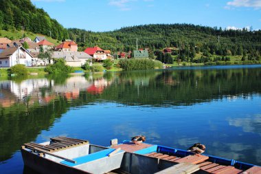 İki yaban ördeği, ormanlık tepelerin, köyün, gölün ve gökyüzünün arka planında yüzen bir teknede oturur..
