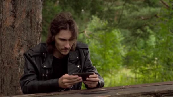 El tipo está sentado con el teléfono en el bosque — Vídeo de stock