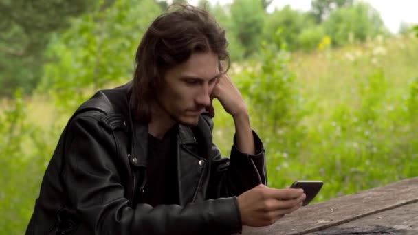 El tipo está sentado con el teléfono en el bosque — Vídeos de Stock