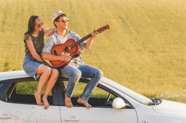 Akustik gitar doğada araba çatıda oturan genç hippi çift. Aşk ve seyahat kavramları.