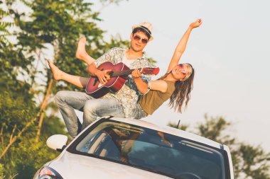 Akustik gitar doğada araba çatıda oturan genç hippi çift. Aşk ve seyahat kavramları.