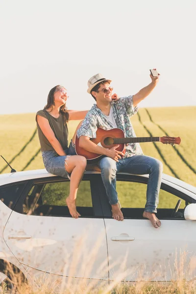 Genç hippi çift araba üzerinde otururken akustik gitar alarak selfie ile doğada çatı.