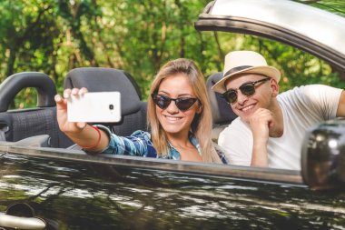 Genç alarak selfie Cabrio arabada otururken smartphone ile çift. Seyahat ve teknoloji kavramları.