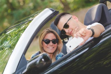 Genç alarak selfie Cabrio arabada otururken smartphone ile çift. Seyahat ve teknoloji kavramları.