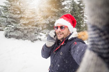 Doğada selfie çekerken smartphone kullanarak Noel Baba şapkası ile genç adam portresi. Kış sahne.