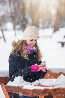Parkta bankta oturmuş ve akıllı telefon kış manzara kullanarak güzel kadın.