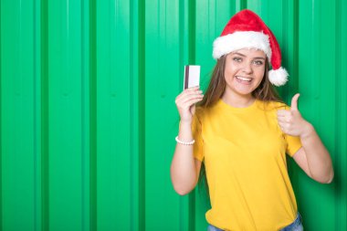 Kredi kartı tutan ve başparmak yeşil arka plan üzerinde gösterilen Noel Baba şapkası olan güzel kadın. Noel alışveriş kavramı. Kopya için alan.