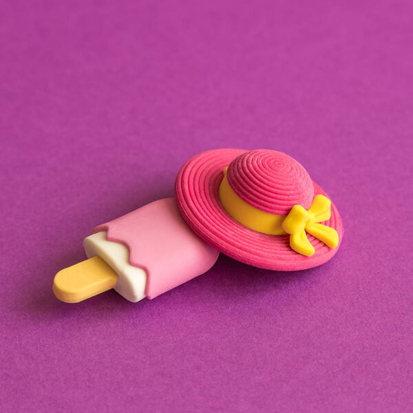 Small ice cream on stick and fashionable sun hat toys against purple background.